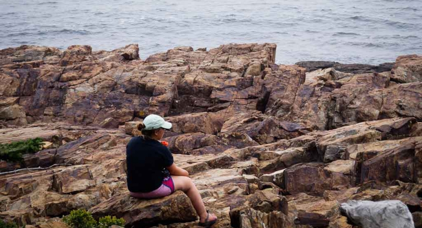 A person sits on a rocky shore, looking out over the water. 